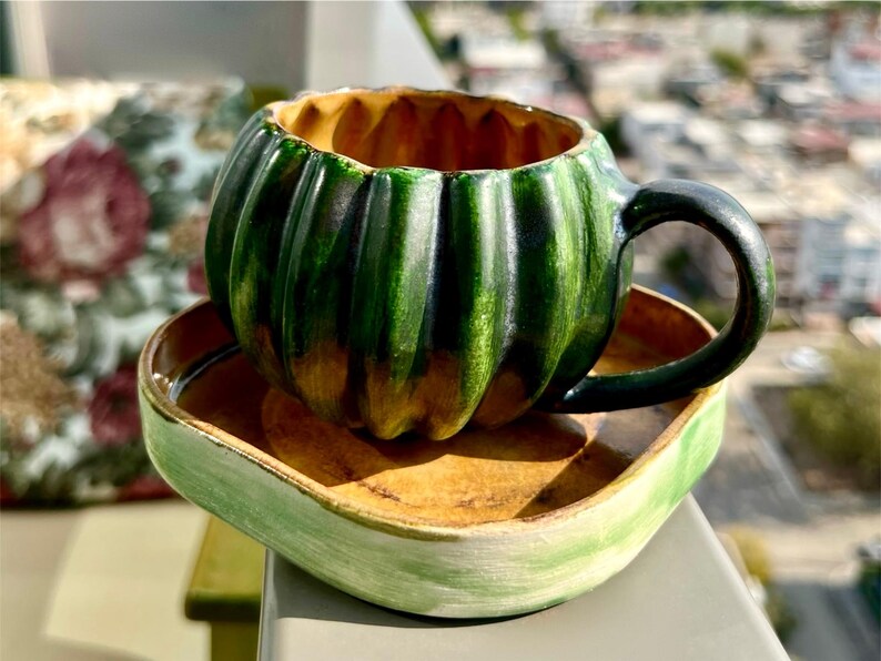 May include: A ceramic teacup and saucer set. The teacup is shaped like a green pumpkin with a black handle. The saucer is square with a light green rim and a brown interior. The set is on a table with a blurred background.