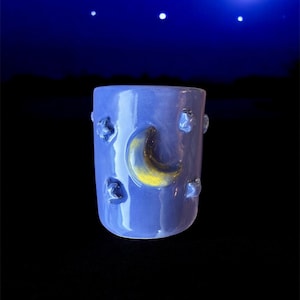Può includere: Una tazza di ceramica blu con un design a mezzaluna e stelle. La tazza ha una finitura lucida e una forma cilindrica. La luna è gialla e le stelle sono in rilievo.