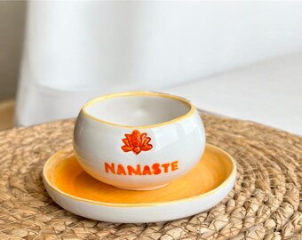 May include: White ceramic bowl with a yellow rim and a small orange lotus flower design. The bowl has the word "NAMASTE" written in orange on the front. The bowl is sitting on a matching yellow saucer.