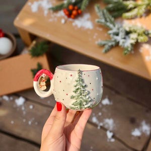 Op de afbeelding: Een witte keramische kerstmok met een groen kerstboomontwerp en rode stippen. De handgreep is rood en heeft een sneeuwpopbedel. De mok staat op een houten oppervlak met decoratief groen.