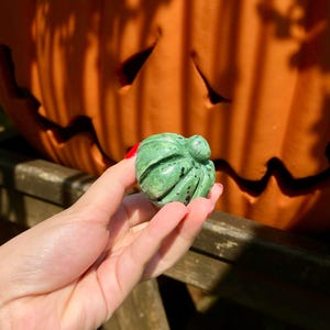 May include: A small, green carved stone pumpkin with dark spots is held in a hand. The pumpkin is in front of a large, carved orange pumpkin. The carved pumpkin has a smiling face. The background is out of focus.