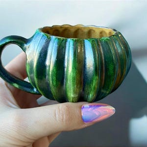 May include: A small, handcrafted ceramic mug with a pumpkin-like shape. The mug features vertical ridges and is glazed in shades of green and blue, with a golden interior. It has a rounded handle and is held by a hand.