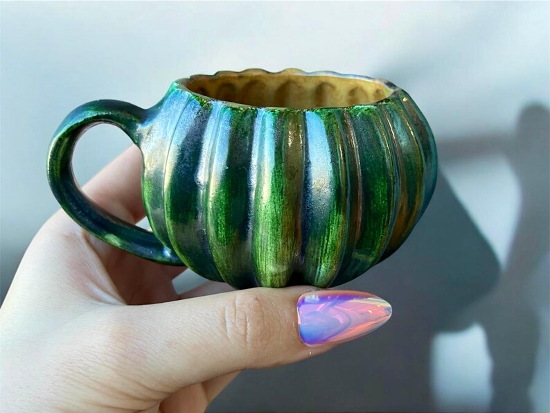 May include: A small, handcrafted ceramic mug with a pumpkin-like shape. The mug features vertical ridges and is glazed in shades of green and blue, with a golden interior. It has a rounded handle and is held by a hand.