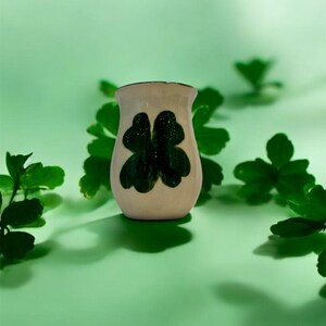 May include: A small, off-white ceramic vase with a dark green four-leaf clover design. The vase is set against a light green background, with green foliage around it. The vase has a slightly flared rim.