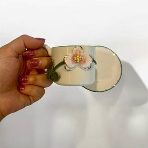 Op de afbeelding: Crèmekleurige kop en schotel set met een handgeschilderd orchidee ontwerp. De orchidee heeft roze en gele details, met groene accenten op de stengel en rand. De kop wordt vastgehouden door een hand met rode glitter nagellak.