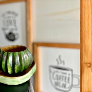May include: A green ceramic mug shaped like a pumpkin, with a matching saucer. The mug has a ribbed design and a brown interior. The saucer is a light green color. In the background are framed pictures.