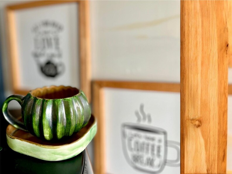 May include: A green ceramic mug shaped like a pumpkin, with a matching saucer. The mug has a ribbed design and a brown interior. The saucer is a light green color. In the background are framed pictures.