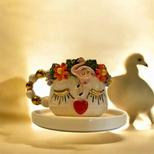 Op de afbeelding: Witte keramische theekop met een gezichtsontwerp, gouden wimpers, een rood hart en een flamingo- en bloemenkroon. Het handvat is versierd met gouden accenten en staat op een bijpassende schotel.
