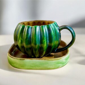 May include: A ceramic teacup and saucer set. The cup is shaped like a pumpkin with vertical ridges and a green glaze. The saucer is square with a brown center and a green rim. The set is designed for serving tea or coffee.