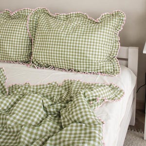 Peut inclure: Un lit avec un drap blanc, deux oreillers à carreaux verts et blancs avec des bords roses à volants, et une housse de couette assortie. La literie est disposée sur un cadre de lit blanc.