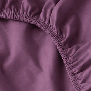May include: Close-up of a fitted sheet in a muted purple color. The elasticized edge is visible, showing the gathered fabric. The sheet appears to be made of a soft, smooth material, suitable for bedding.