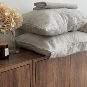 Può includere: Una pila di cuscini in lino grigio chiaro e un lenzuolo piegato su un mobile in legno. Un vaso di vetro trasparente con fiori secchi e una candela in un barattolo di vetro sono sul mobile. I cuscini hanno un aspetto strutturato.