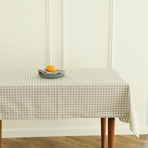 Puede incluir: Un mantel rectangular con un patrón de cuadros grises y blancos cubre una mesa de madera. Una pila de platos grises, una servilleta gris y una fruta naranja están sobre la mesa. El fondo es una pared blanca.