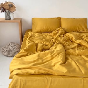 Könnte beinhalten: Ein Bett mit einem senfgelben Bettwäsche-Set, einschließlich eines Spannbetttuchs, eines Bettbezugs und zwei Kissenbezügen. Die Bettwäsche ist leicht zerknittert, was auf Komfort hindeutet. Ein hölzerner Beistelltisch mit einer Vase mit Trockenblumen und ein kleiner, runder Pouf im Hintergrund.