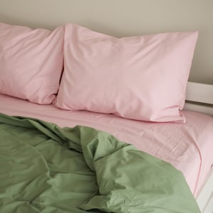 May include: A bed is made with a light pink fitted sheet and two matching pillowcases. A green comforter is partially visible, creating a contrast. The bedding is neatly arranged, suggesting a clean and inviting bedroom setting.