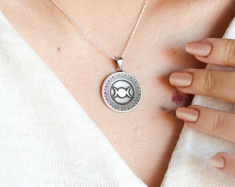 May include: A silver necklace with a round pendant featuring a triple moon symbol surrounded by a Greek key design.