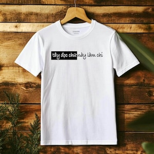 May include: A white t-shirt with a crew neck, hanging on a wooden hanger. The shirt features black text in a rectangular box. The background is a wooden surface with green foliage on the left and right sides.