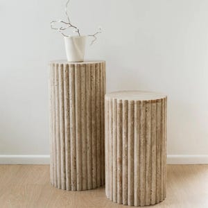 May include: Two fluted cylindrical pedestals made of travertine stone. The pedestals are different sizes and are stacked on top of each other. The larger pedestal is topped with a white ceramic vase.