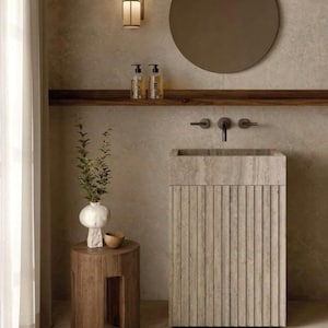 May include: A modern bathroom vanity with a stone sink and a wooden shelf. The vanity has a textured, light grey finish and a round mirror above it. There is a small wooden stool with a vase and a bowl on top.