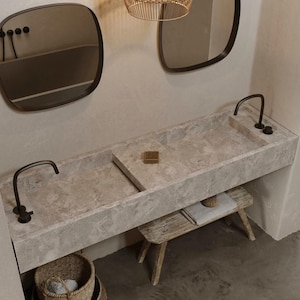 Peut inclure: Une vasque de salle de bain double avec un plan de travail en pierre gris clair et deux robinets noirs. Le meuble est monté sur un mur et possède une étagère en bois en dessous. Il y a un petit savon dans l'évier à droite.