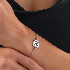 Pode incluir: Uma pulseira de prata com um pingente quadrado geométrico com um design floral vazado. A pulseira é delicada e tem uma corrente fina. A joia é usada em um pulso contra um fundo preto.
