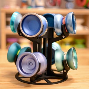 May include: A black, multi-tiered yo-yo display stand holding several colorful yo-yos. The yo-yos are various shades of blue, teal, and purple, with silver rims. The stand is on a wooden surface, with a blurred background.