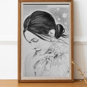 May include: A framed pencil sketch of a woman with her hair in a bun, gazing downwards. The artwork is in monochrome, with delicate shading and detail. The frame is a warm brown, and the artwork is displayed on a wooden surface.