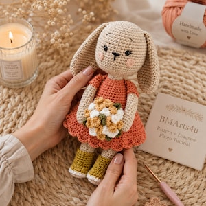 May include: A beige crocheted rabbit doll wearing a coral-colored dress with a scalloped hem and yellow boots. The rabbit holds a small bouquet of white and yellow flowers. A card with the text BMArts4u Pattern + Diagram Photo Tutorial is visible.