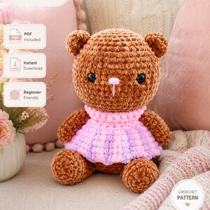 May include: A brown crocheted teddy bear plush toy wearing a pink and light purple striped dress. The bear has black plastic eyes and a small pink nose. The image includes text labels indicating PDF included, instant download, and beginner friendly.