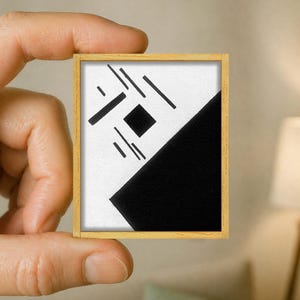 Op de afbeelding: Een klein rechthoekig kunstwerk in een lichthouten lijst met een witte achtergrond, een zwarte driehoek in de rechterbenedenhoek en een reeks zwarte lijnen en een enkel zwart vierkant in het linkerbovenste gedeelte.