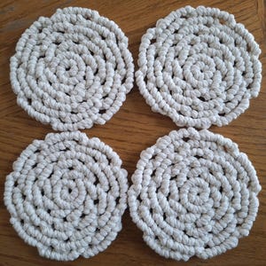 May include: Four off-white, circular coasters made from tightly woven rope. The coasters have a spiral design and are arranged on a wooden surface. The coasters are a neutral color and would complement any decor.