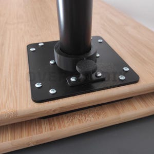 Könnte beinhalten: Nahaufnahme eines schwarzen Metall-Tischbein-Sockels, der an einer hellbraunen Bambusoberfläche befestigt ist. Der quadratische Sockel hat abgerundete Ecken und ist mit silbernen Schrauben befestigt. Ein schwarzes zylindrisches Bein erhebt sich aus der Mitte.