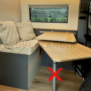 Op de afbeelding: Een houten tafel met een neerklappbare verlenging, gemonteerd aan de muur in een camper. De tafel is gemaakt van licht hout en heeft een grijs metalen steunpoot. Een rode X is over de poot getekend.