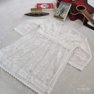 May include: White cotton tunic with embroidered floral details and a lace trim. The tunic has a V-neckline and three-quarter sleeves.