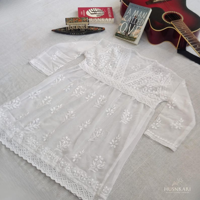 May include: White cotton tunic with embroidered floral details and a lace trim. The tunic has a V-neckline and three-quarter sleeves.