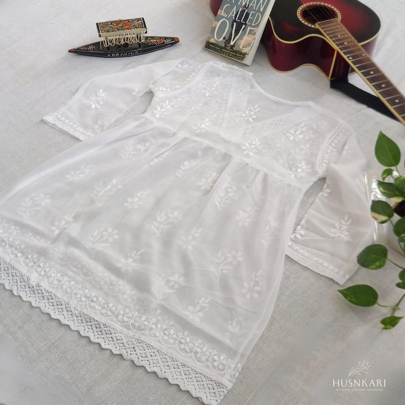 May include: White cotton tunic with embroidered floral details and a lace trim. The tunic has a V-neckline and 3/4 sleeves.