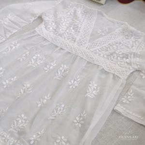 May include: White cotton dress with a lace trim and floral embroidery. The dress has a V-neckline and long sleeves.