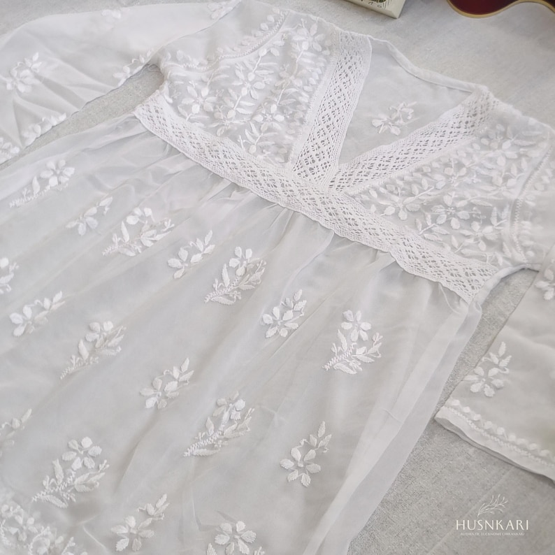 May include: White cotton dress with a lace trim and floral embroidery. The dress has a V-neckline and long sleeves.