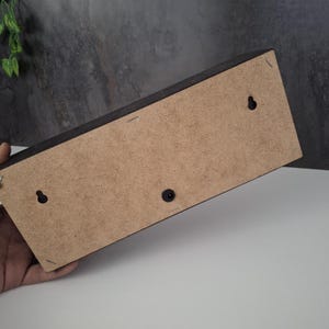 May include: A rectangular wooden box with a dark brown exterior and a light brown back. The back has two keyhole hangers and a black plastic button. The box is held by a hand.