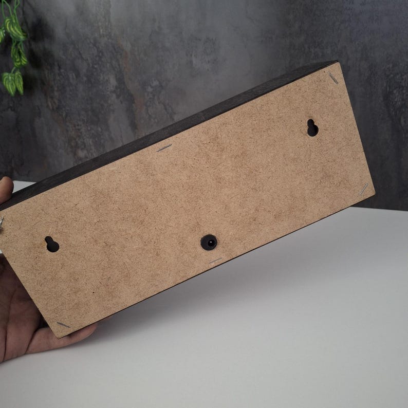 May include: A rectangular wooden box with a dark brown exterior and a light brown back. The back has two keyhole hangers and a black plastic button. The box is held by a hand.