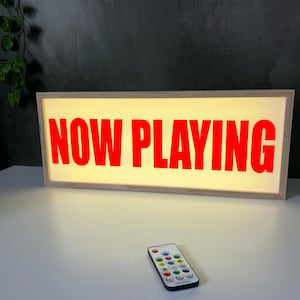 May include: Illuminated sign with the text "NOW PLAYING" in red. The sign is in a wooden frame, and a remote control is placed in front of it. The sign is rectangular.