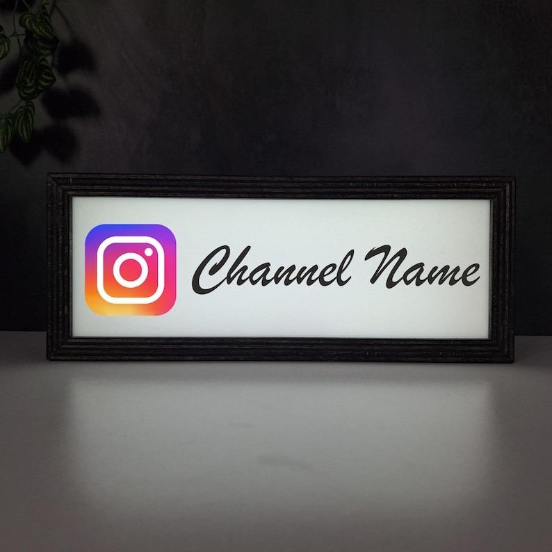 May include: A rectangular, black-framed sign with a white background. The Instagram logo is on the left, with a gradient of orange, pink, and purple. The words "Channel Name" are written in cursive on the right.