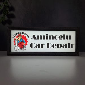 May include: Rectangular black framed sign with the text "Aminoglu Car Repair". The sign also features a cartoon mechanic leaning against a wrench and a gear with the words "Service Center".