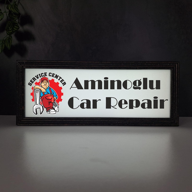 May include: Rectangular black framed sign with the text "Aminoglu Car Repair". The sign also features a cartoon mechanic leaning against a wrench and a gear with the words "Service Center".