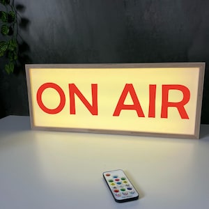 Peut inclure: Un panneau lumineux rectangulaire avec les mots "ON AIR" en lettres rouges. Le panneau a un cadre en bois et un fond jaune vif. Une télécommande avec plusieurs boutons colorés est devant le panneau.