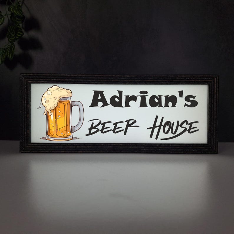 May include: A framed sign with the text "Adrian's BEER House" in black lettering. The sign features a cartoon illustration of a beer mug filled with golden beer and foamy head. The frame is dark brown.