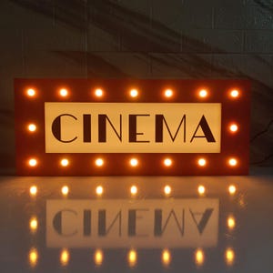 Può includere: Un'insegna rettangolare illuminata con la scritta "CINEMA" in lettere nere. L'insegna è incorniciata in legno marrone scuro e delineata da lampadine a luce calda, distanziate in modo uniforme. Il riflesso dell'insegna è visibile sotto.