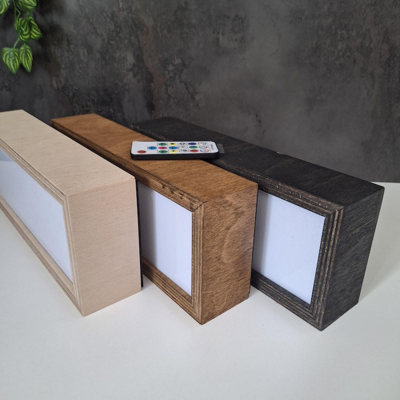 May include: Three rectangular wooden LED light boxes in natural, medium brown, and dark brown finishes. A remote control rests on the brown box. Each box has a white illuminated interior.