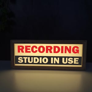 以下が含まれることがあります： 長方形の木製フレームの看板で、赤い「RECORDING」と白い「STUDIO IN USE」の文字が表示されています。看板は点灯しており、スタジオが使用中であることを示しています。