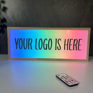 May include: Rectangular light box with a wooden frame, displaying the text "YOUR LOGO IS HERE" in black. The background is a vibrant gradient of rainbow colors. A remote control is in the foreground.